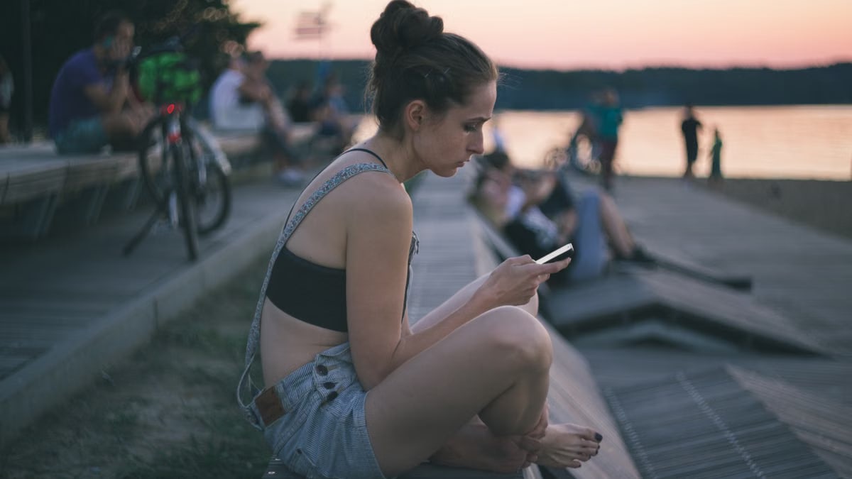 A young woman sits by the lakeside using her smartphone, enjoying a tranquil sunset.